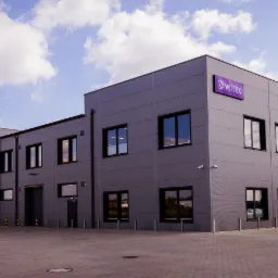 Szary budynek firmy odzieżowej Wiltrex z fioletowym logo na rogu, widoczny z zewnątrz na tle błękitnego nieba z chmurami.