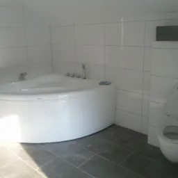 Biała narożna wanna z hydromasażem i toaleta w nowoczesnej łazience wyłożonej białymi płytkami, szara podłoga.