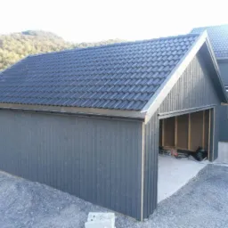 Nowoczesny garaż z ciemnoszarą elewacją i grafitowym dachem z dach&oacute;wki ceramicznej, widok na wnętrze w stanie surowym, z betonową posadzką i deskami na ścianach, na tle wzg&oacute;rz.