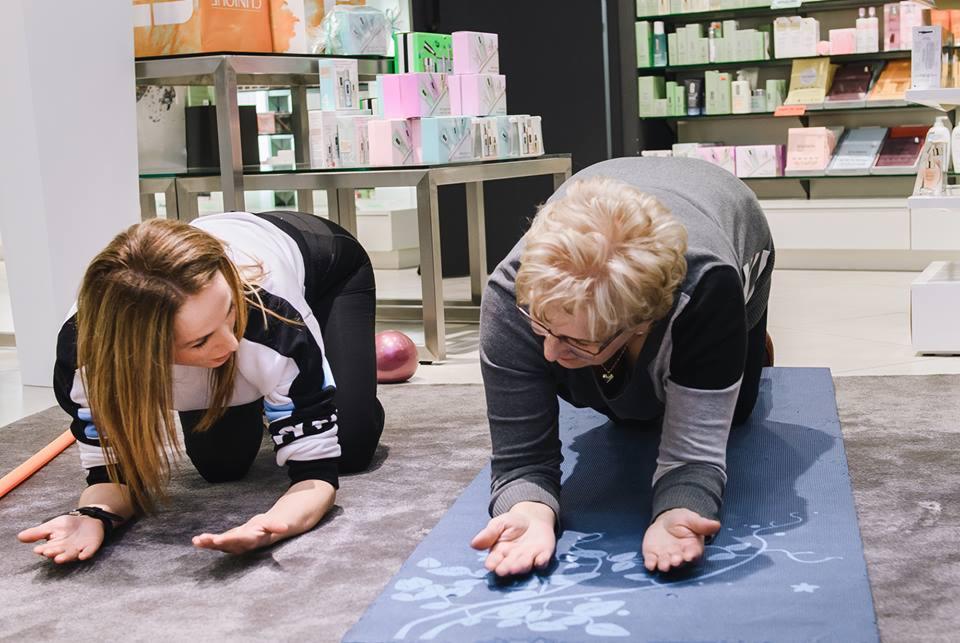 Dwie kobiety w pozycji deski na kolanach i dłoniach, ćwiczące na macie do jogi w jasnym pomieszczeniu, w tle kosmetyki na półkach.