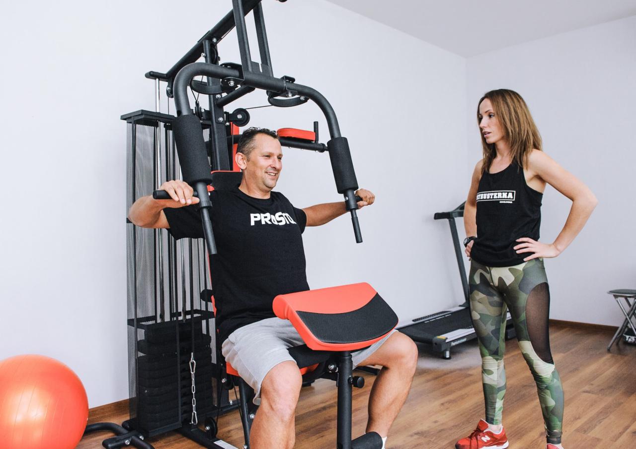 Mężczyzna ćwiczy na atlasie pod okiem kobiety w sportowym stroju, widoczna bieżnia w tle.