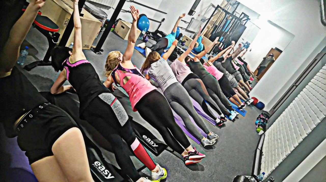 Grupa osób w sportowych strojach wykonuje ćwiczenia rozciągające na matach w jasnej sali fitness, z uniesionymi rękami i widocznym logo 'easy fitness' na sprzęcie.