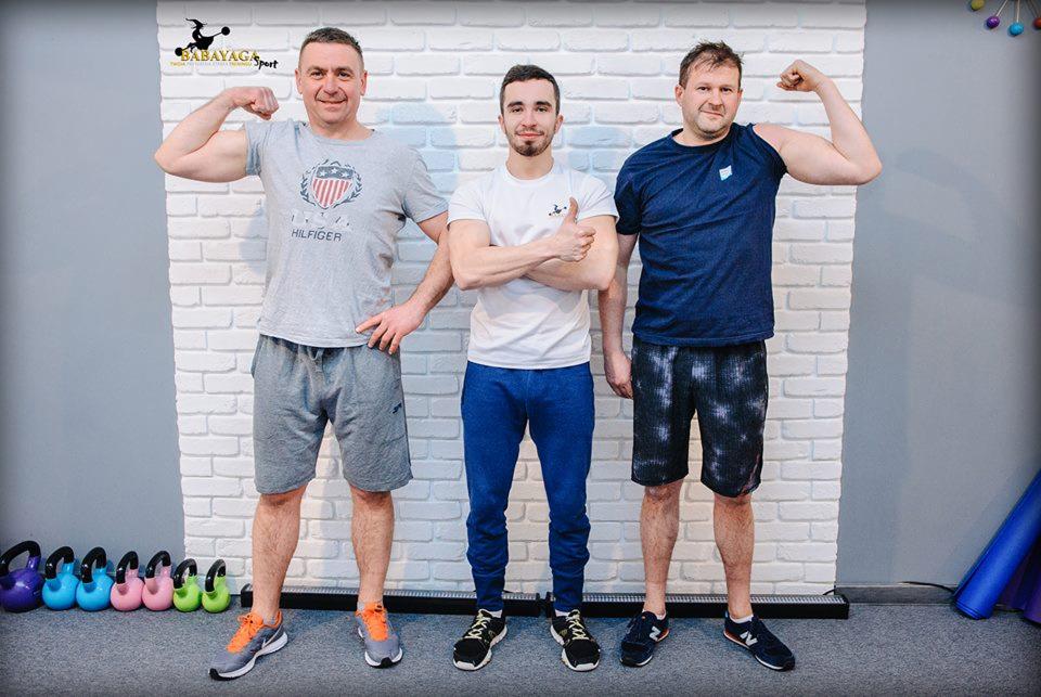 Trzech mężczyzn pozuje na tle białej ceglanej ściany w siłowni, prezentując bicepsy i uśmiechając się; w tle widoczne kolorowe kettlebells.