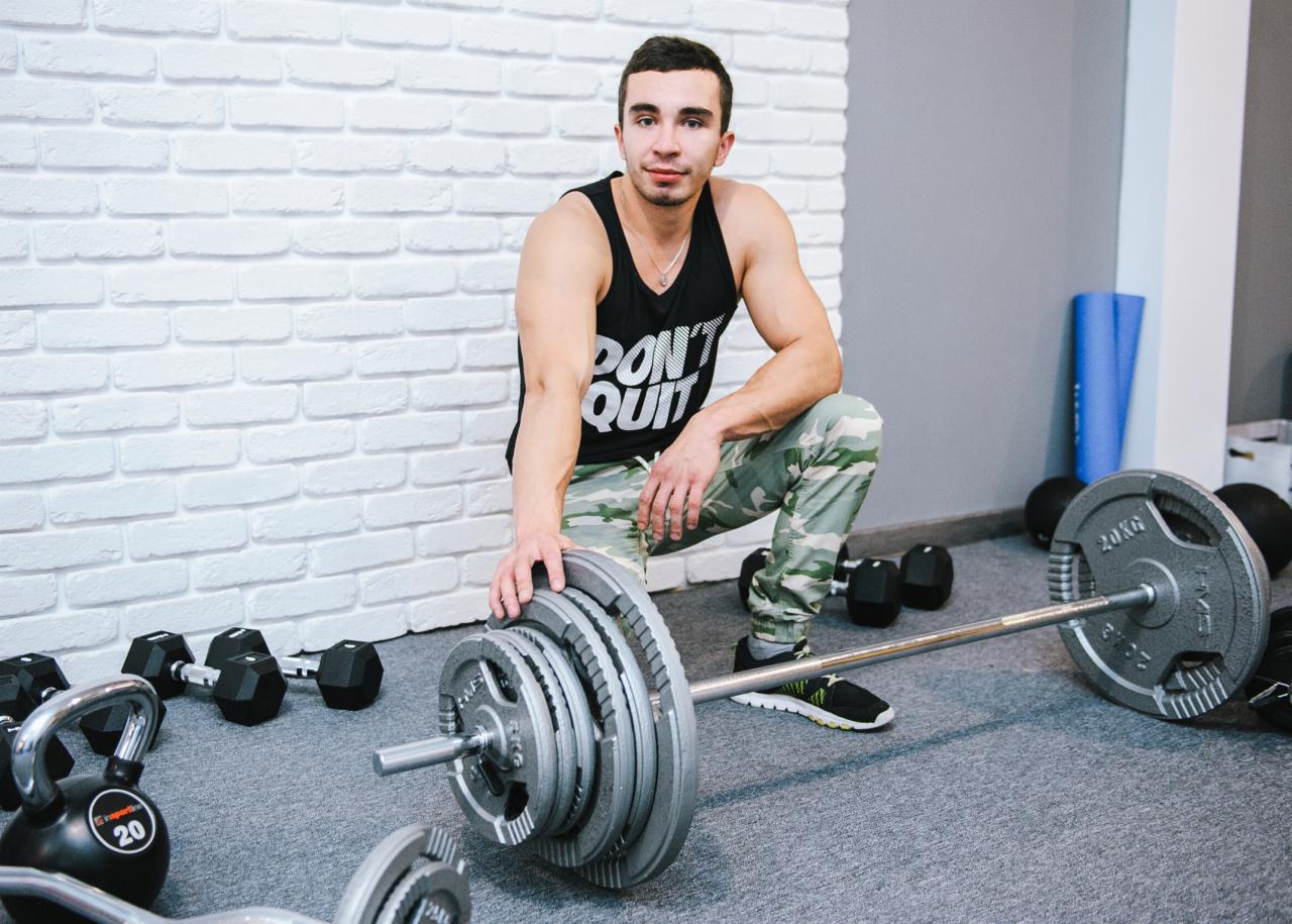 Mężczyzna w sportowym stroju kuca obok sztangi z obciążeniami w siłowni, ubrany w czarny tank top z napisem 'DON'T QUIT' i spodnie moro.