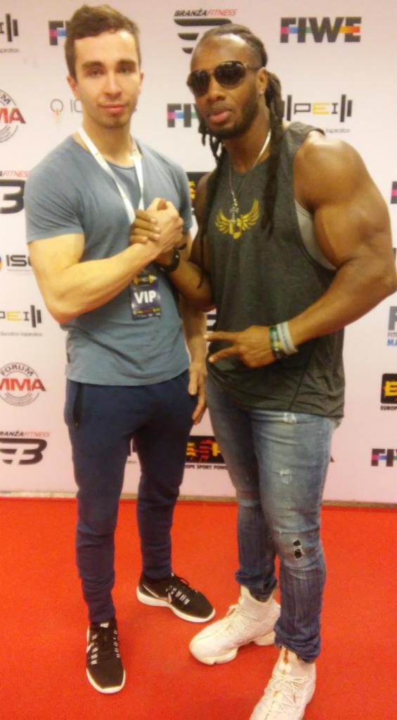 Dwóch mężczyzn, jeden z widoczną muskulaturą, w okularach przeciwsłonecznych, pozuje na tle ściany z logo branży fitness, drugi z identyfikatorem VIP, ściskają sobie dłonie.