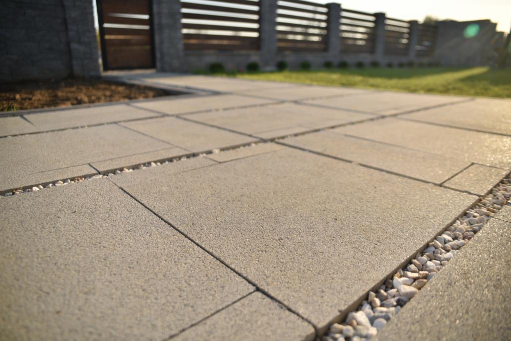 Nowoczesna nawierzchnia z szarych płyt betonowych, wypełniona białym grysem, prowadząca do drewnianej bramy z poziomymi szczeblami i betonowego ogrodzenia w tle.