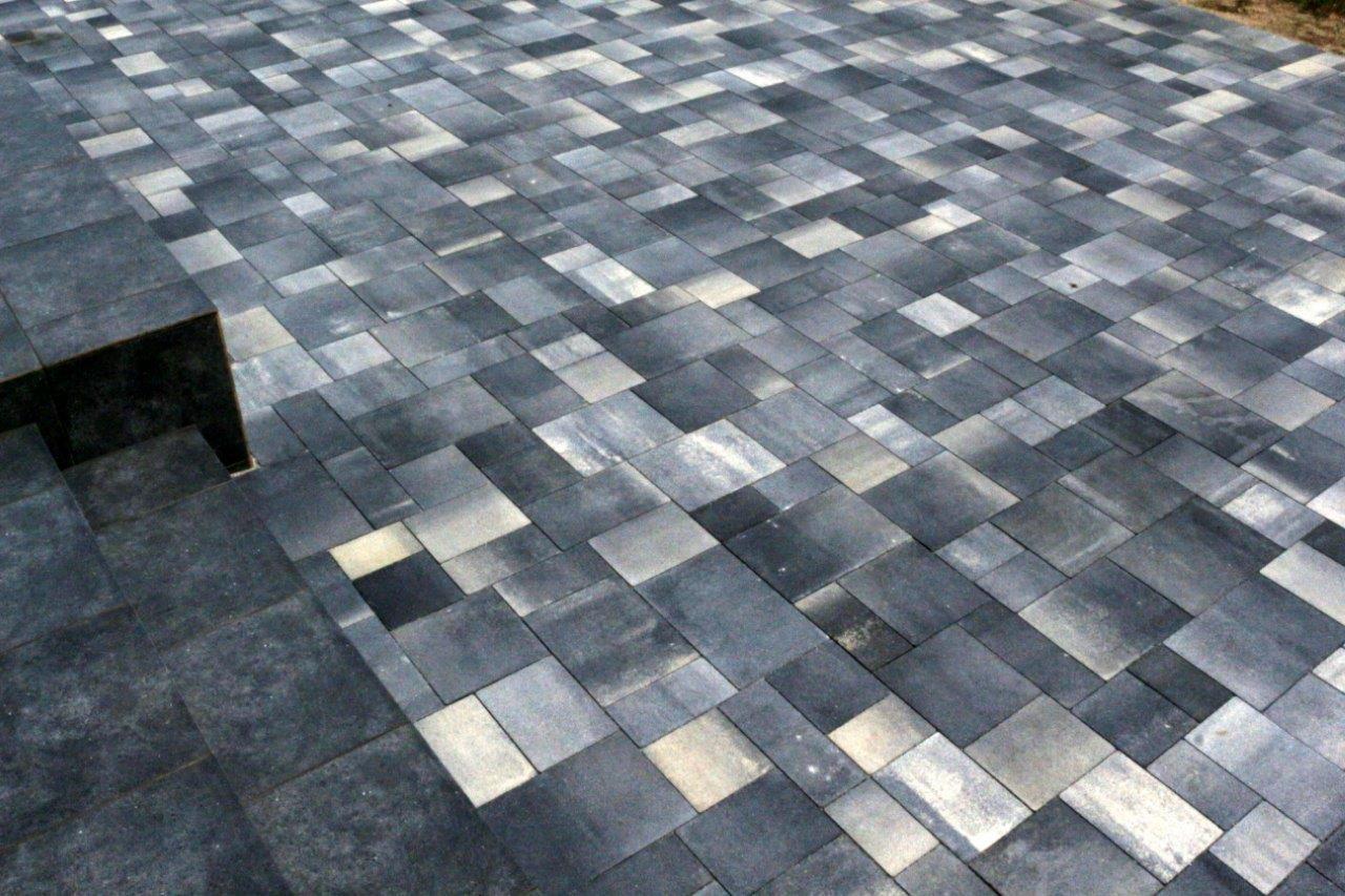 Ułożona kostka brukowa w odcieniach szarości i grafitu, wzór z prostokątnych i kwadratowych elementów, schody z kostki po lewej stronie kadru.