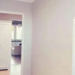 Pomalowany na jasny odcień szarości korytarz z widocznym fragmentem salonu w głębi, ukazujący białe meble i okno z delikatnymi firanami.