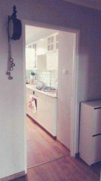 Widok kuchni z białymi szafkami i drewnianą podłogą przez otwarte drzwi, z zegarem na ścianie po lewej stronie i komodą po prawej.