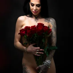 Kobieta z tatuażami pozuje z bukietem czerwonych róż na ciemnym tle, patrząc w dół, z widocznymi detalami biżuterii na dłoniach i dekolcie.