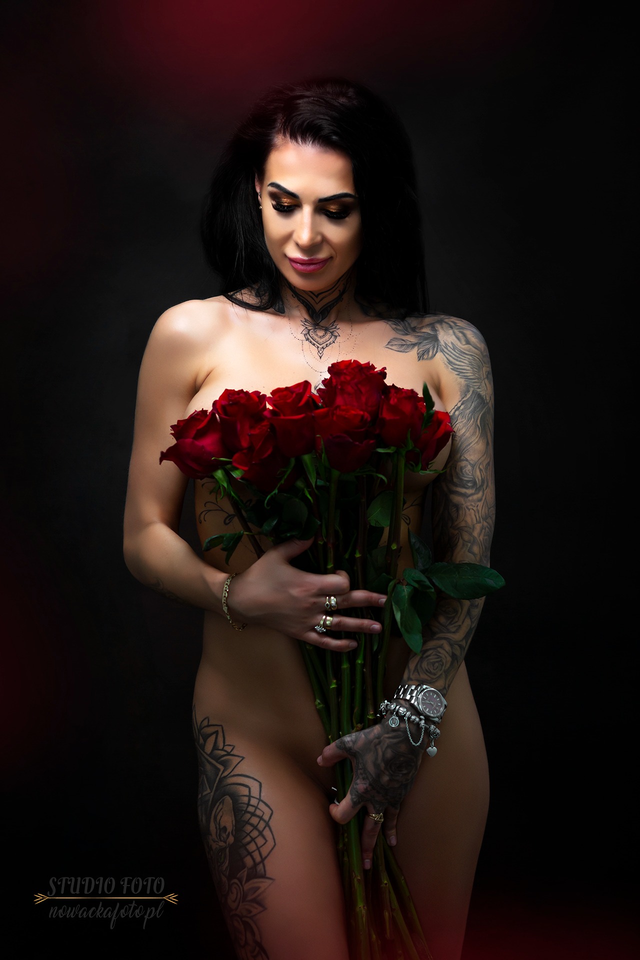 Kobieta z tatuażami pozuje z bukietem czerwonych róż na ciemnym tle, patrząc w dół, z widocznymi detalami biżuterii na dłoniach i dekolcie.