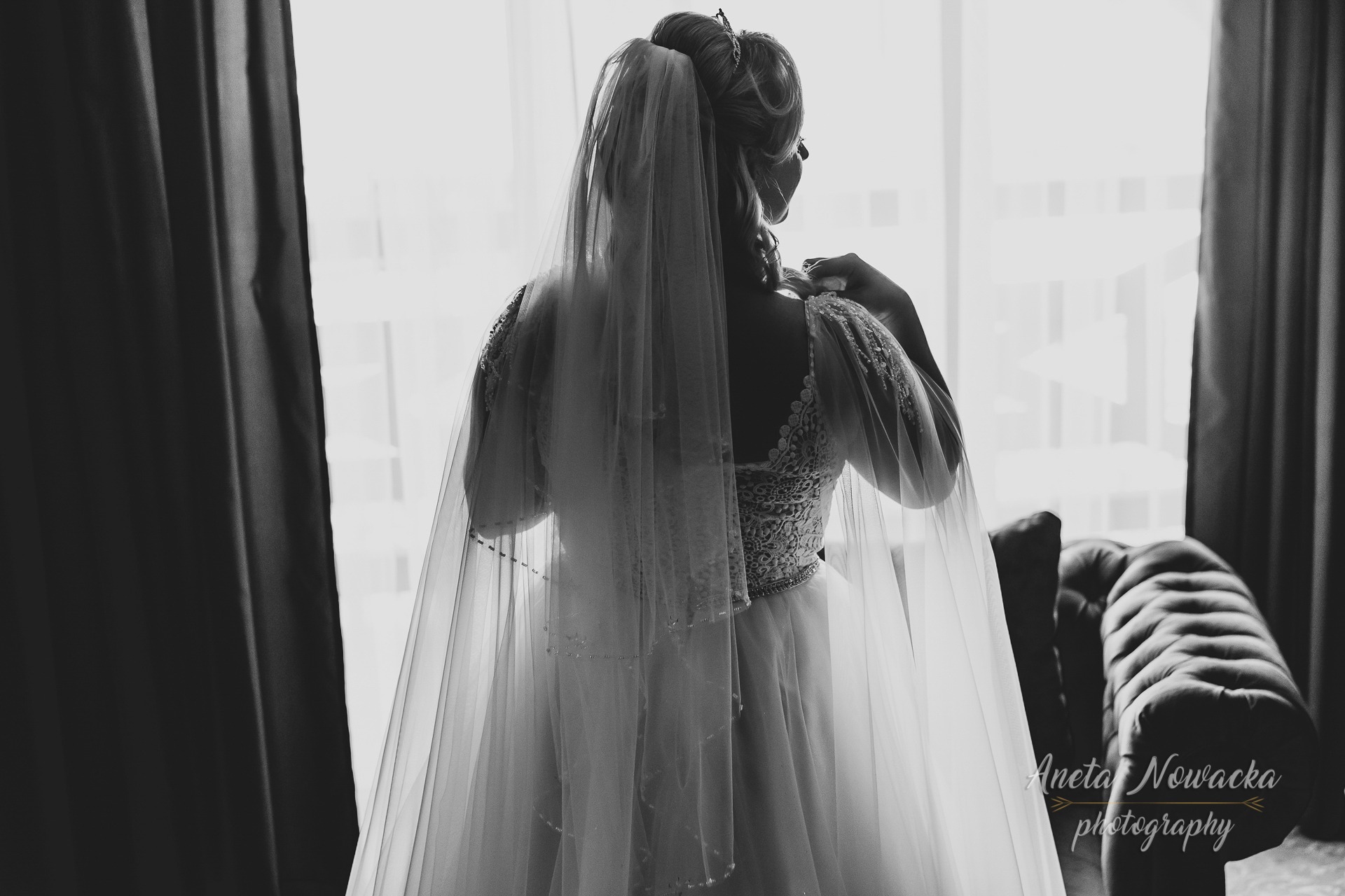 Panna młoda w koronkowej sukni ślubnej i welonie, stojąca tyłem przy oknie z zasłoniętą kurtyną, czarno-białe zdjęcie z widocznym logiem fotografa.