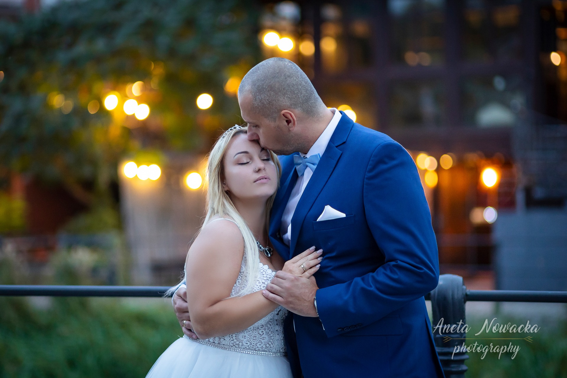 Mężczyzna w niebieskim garniturze całuje w czoło blondynkę w białej sukni ślubnej na tle rozmytych światełek.
