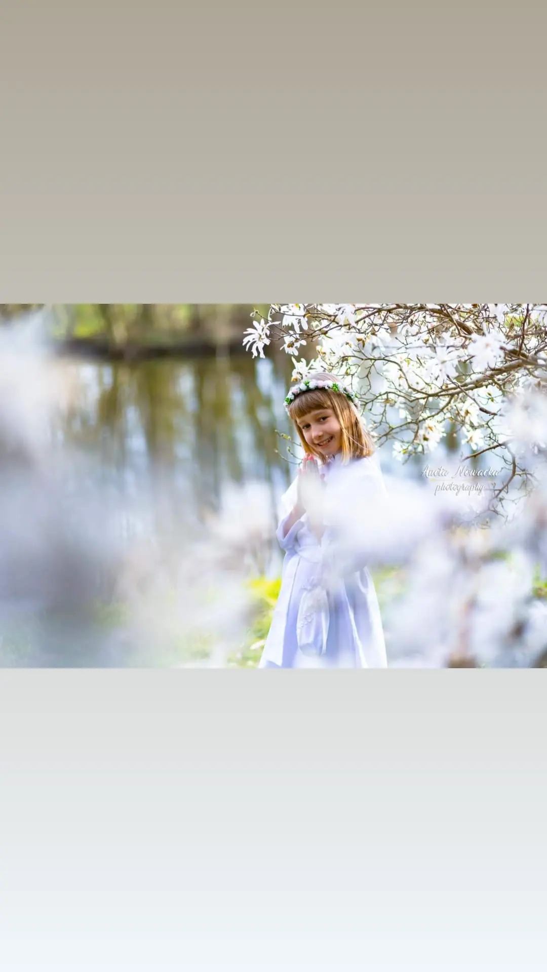 Uśmiechnięta dziewczynka w białej sukience i wianku na głowie, pozuje na tle kwitnącej magnolii i odbijającego się w wodzie krajobrazu.