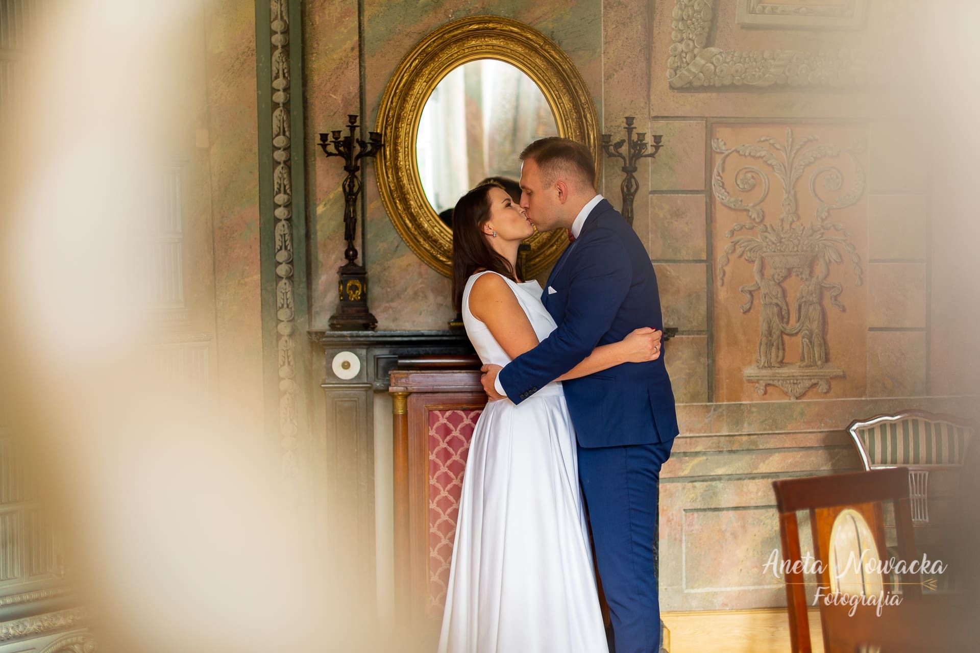 Pocałunek młodej pary na tle lustra w złotej ramie i dekoracyjnej ściany z ornamentami w pałacowym wnętrzu.
