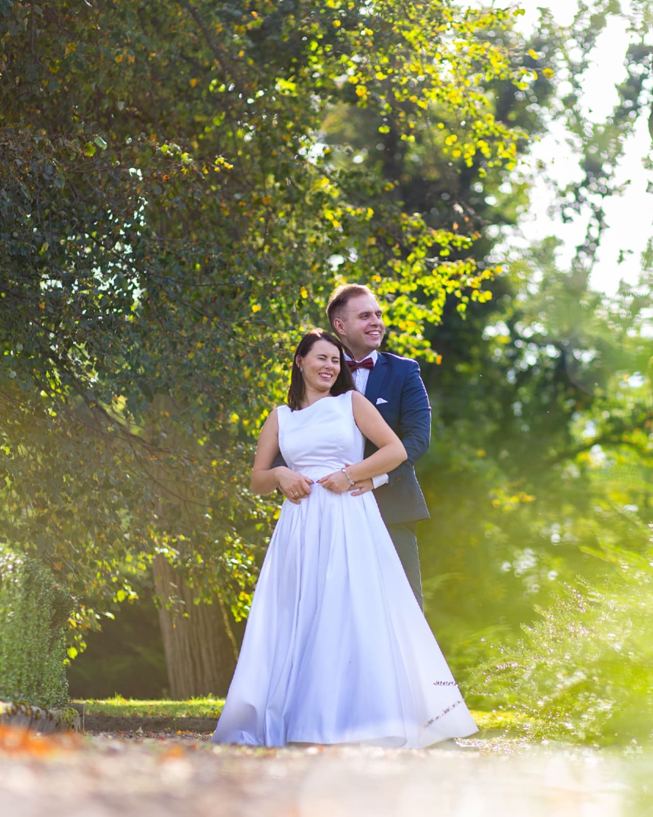 Uśmiechnięta para w plenerze: kobieta w białej sukni z rękami na talii, mężczyzna w granatowym garniturze i bordowej muszce, oboje patrzą w dal na tle drzew i zieleni.