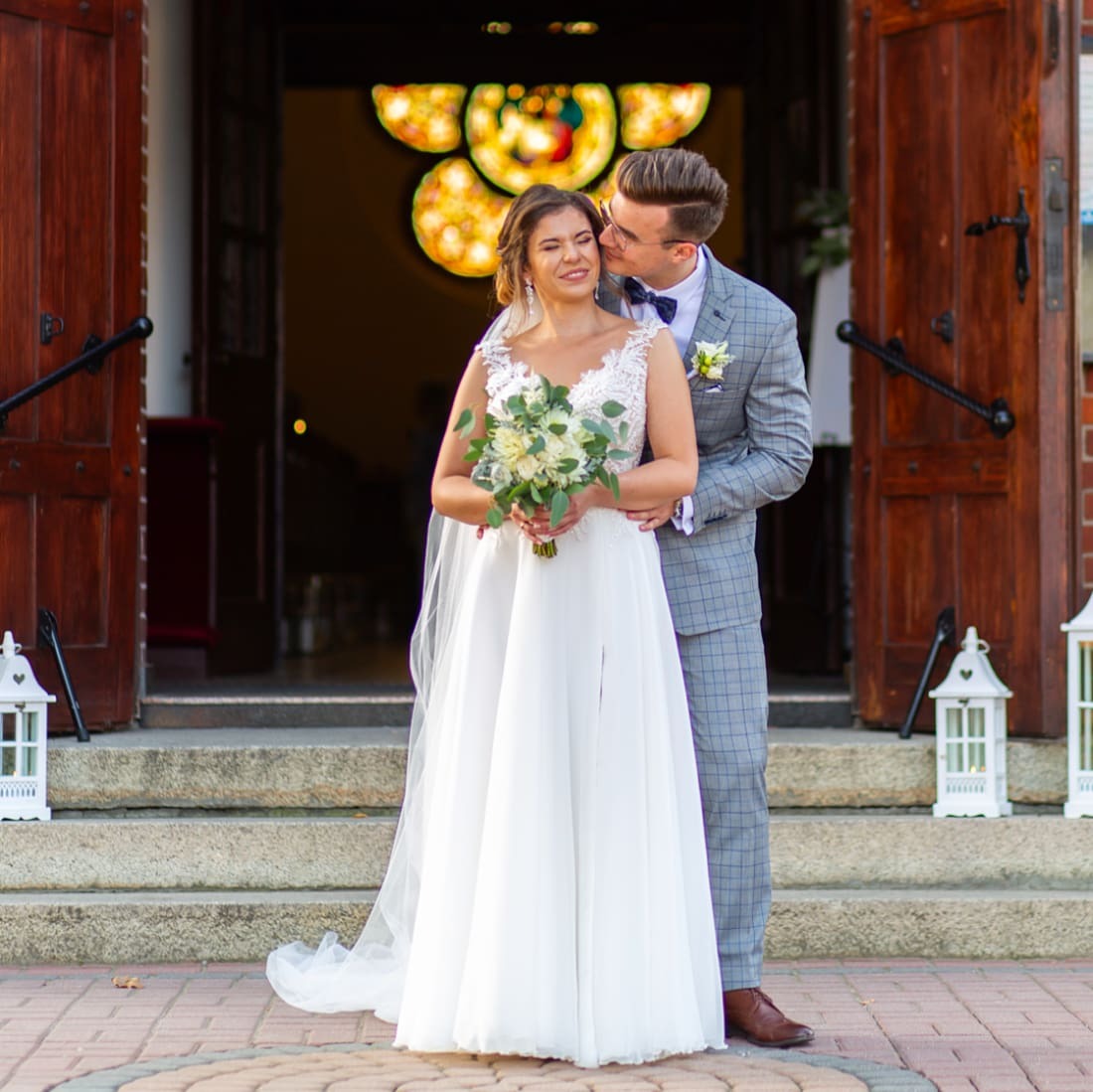 Uśmiechnięta panna młoda w koronkowej sukni z bukietem, całowana w policzek przez pana młodego w szarej kraciastej marynarce, na tle wejścia do kościoła z witrażem.