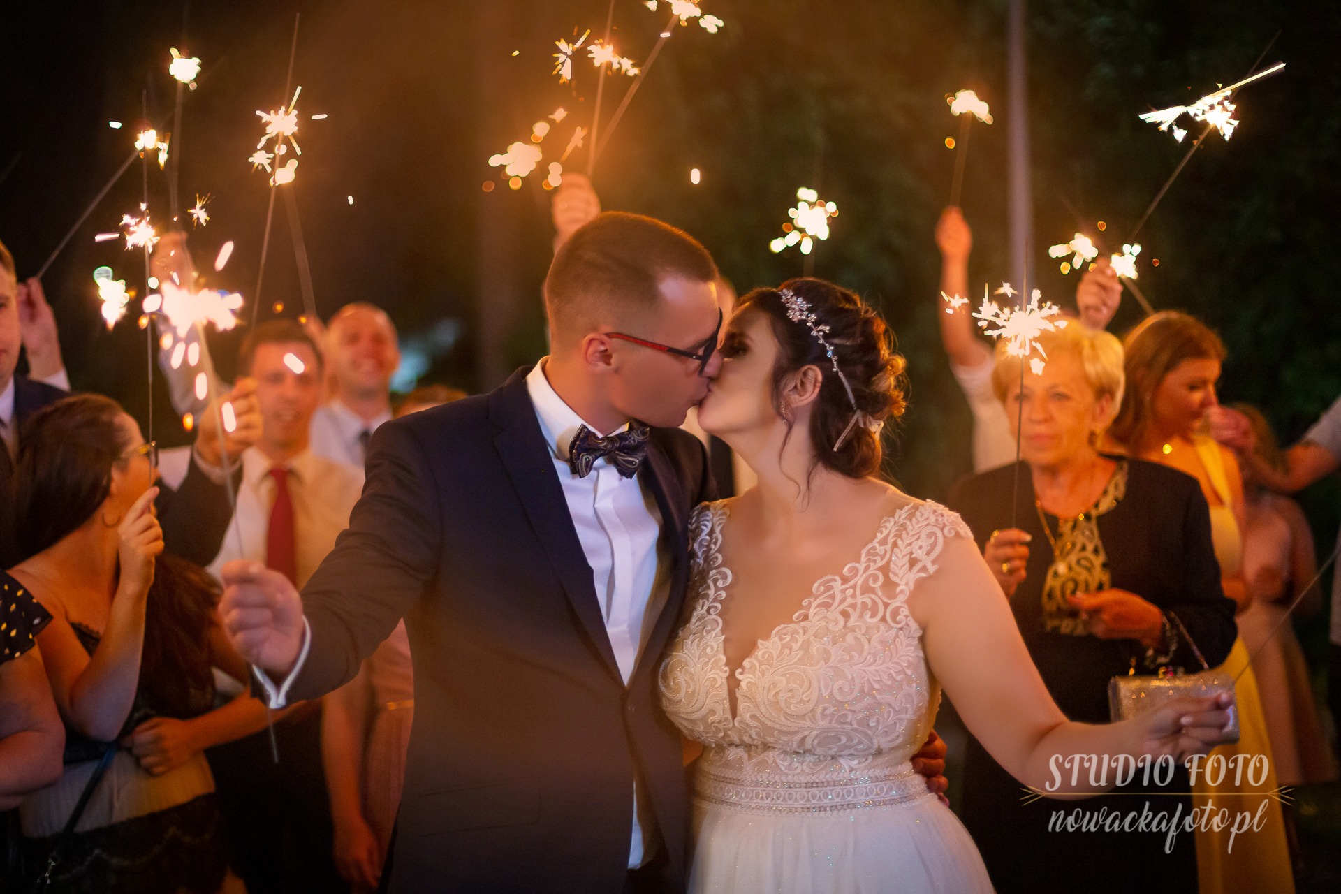 Pocałunek pary młodej otoczonej gośćmi z zimnymi ogniami, panna młoda w koronkowej sukni, pan młody w granatowym garniturze z muszką w paisley, ciemne tło.