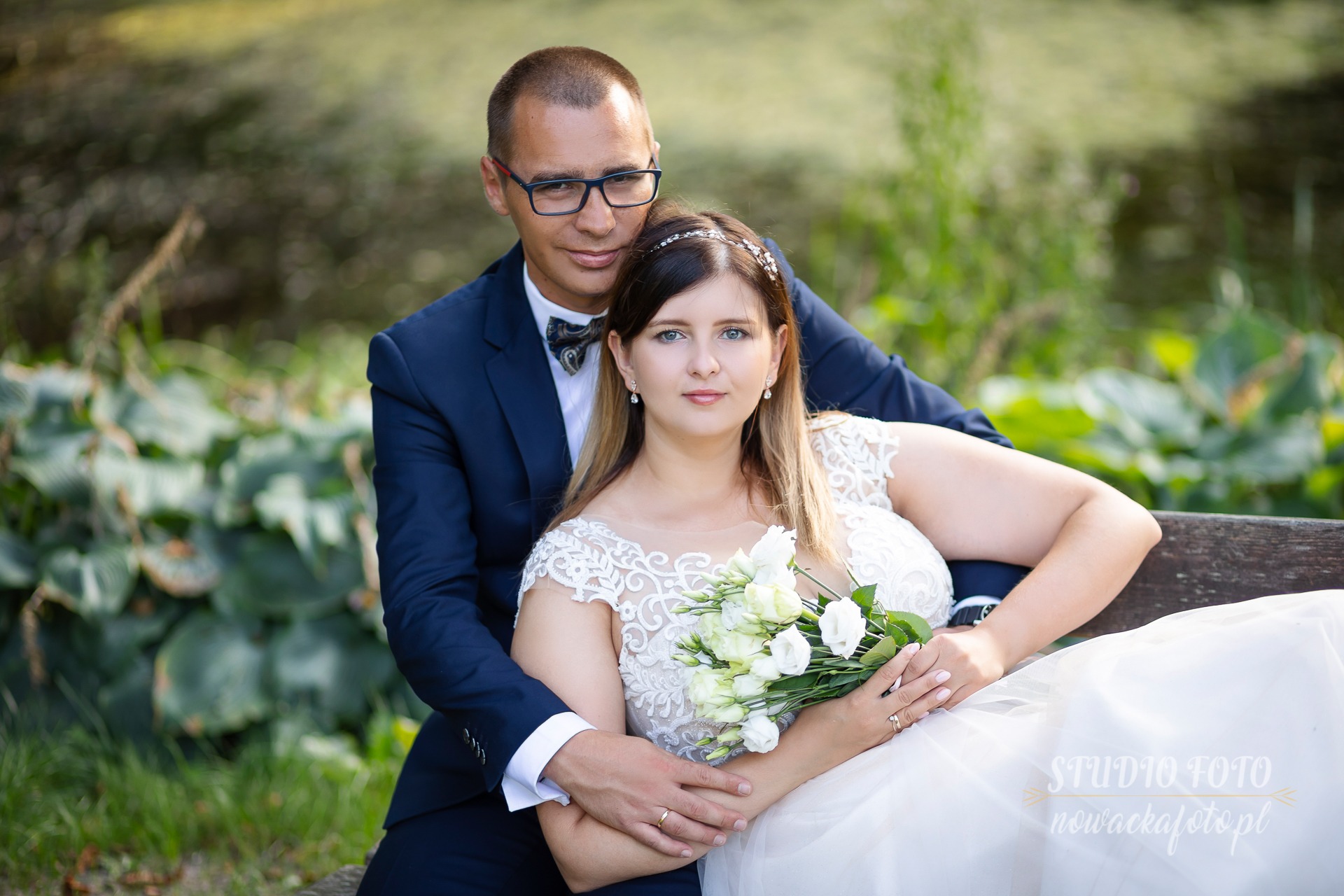 Para młoda siedząca na ławce w parku, panna młoda w białej sukni z koronkową górą trzyma bukiet białych róż, pan młody w granatowym garniturze i muszce obejmuje ją ramionami, w tle zieleń drzew...