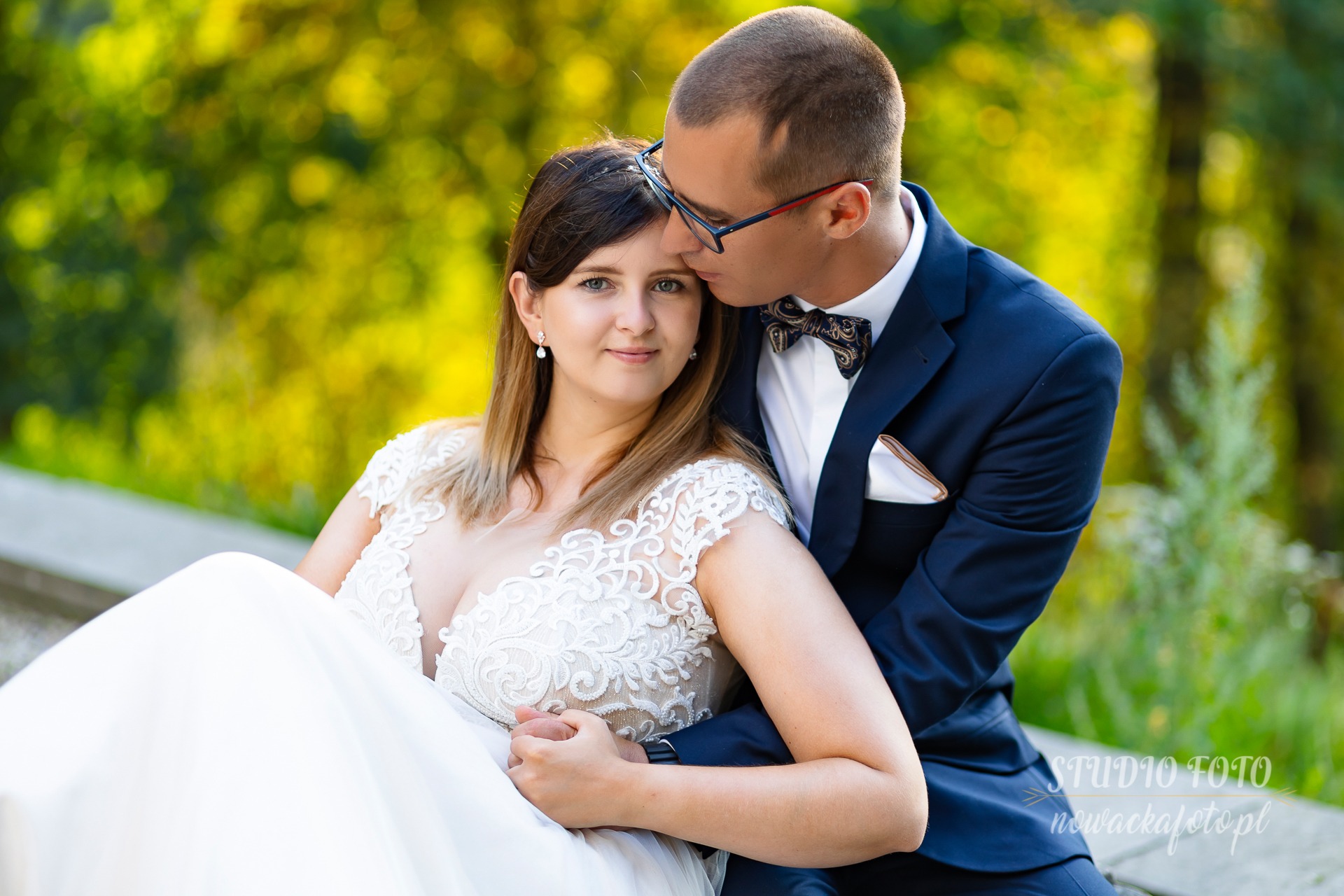 Uśmiechnięta panna młoda w koronkowej sukni, obejmowana przez pana młodego w granatowym garniturze z muszką, delikatny pocałunek w czoło na tle zieleni.