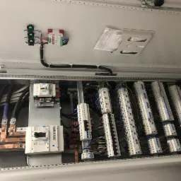 Otwarta szafa elektryczna z widocznymi przewodami, listwami zaciskowymi i zabezpieczeniami, zainstalowana wewnątrz pomieszczenia.