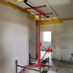 Montaż płyty gipsowo-kartonowej na suficie za pomocą podnośnika, widoczne żółte taśmy uszczelniające na łączeniach, surowy stan wykończenia wnętrza.