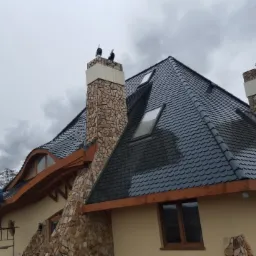 Wykończony dach z ciemnej dachówki na budynku z kamiennymi kominami i drewnianymi elementami. Pracownik na rusztowaniu przy krawędzi dachu.