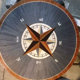 Okrągły blat drewniany z intarsją w kształcie róży wiatrów, widoczne kierunki świata, wykończony brązową obwódką, w tle warsztat stolarski.