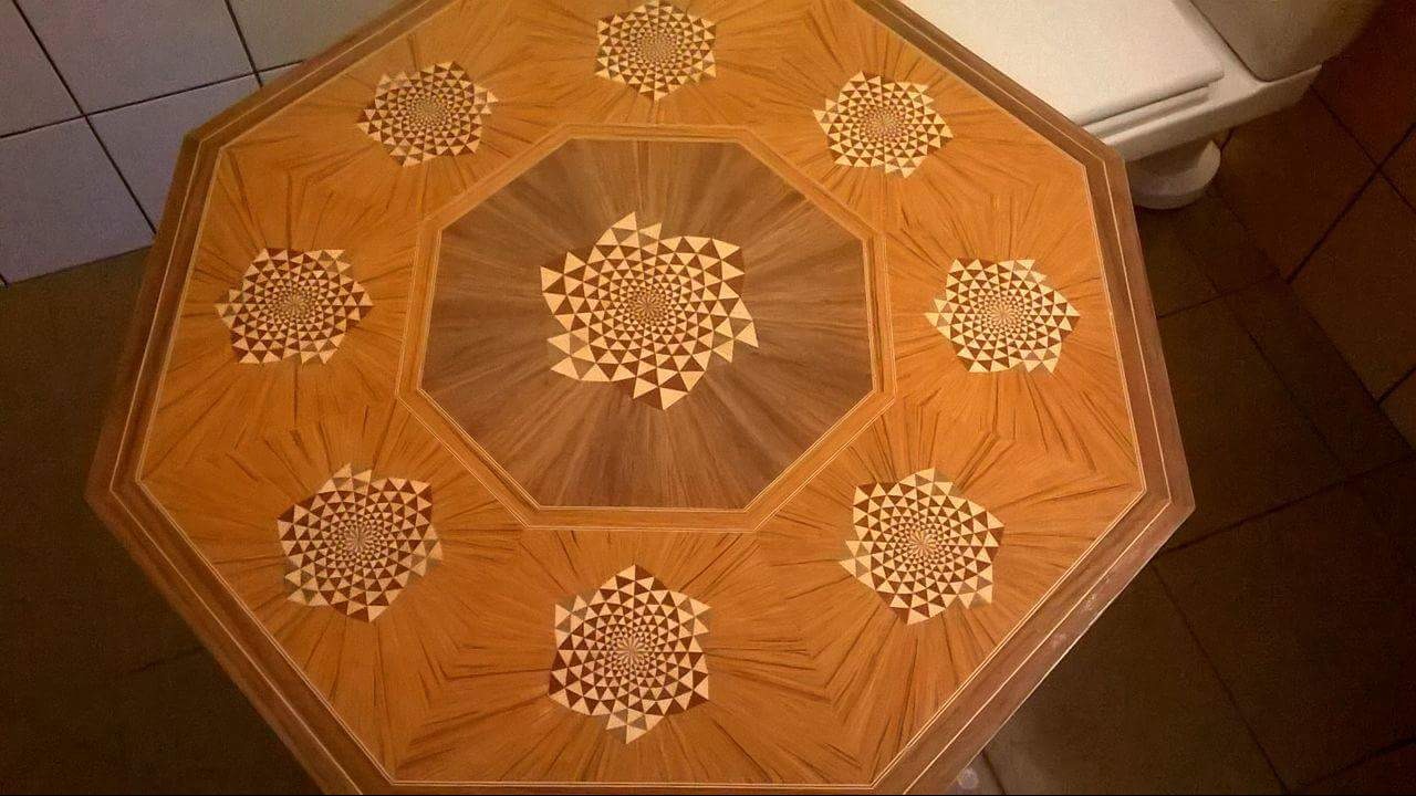 Ośmiokątny blat stołu z intarsją z różnych gatunków drewna, tworzącą geometryczne wzory w kształcie rozet z trójkątów, widok z góry, ciepłe oświetlenie.