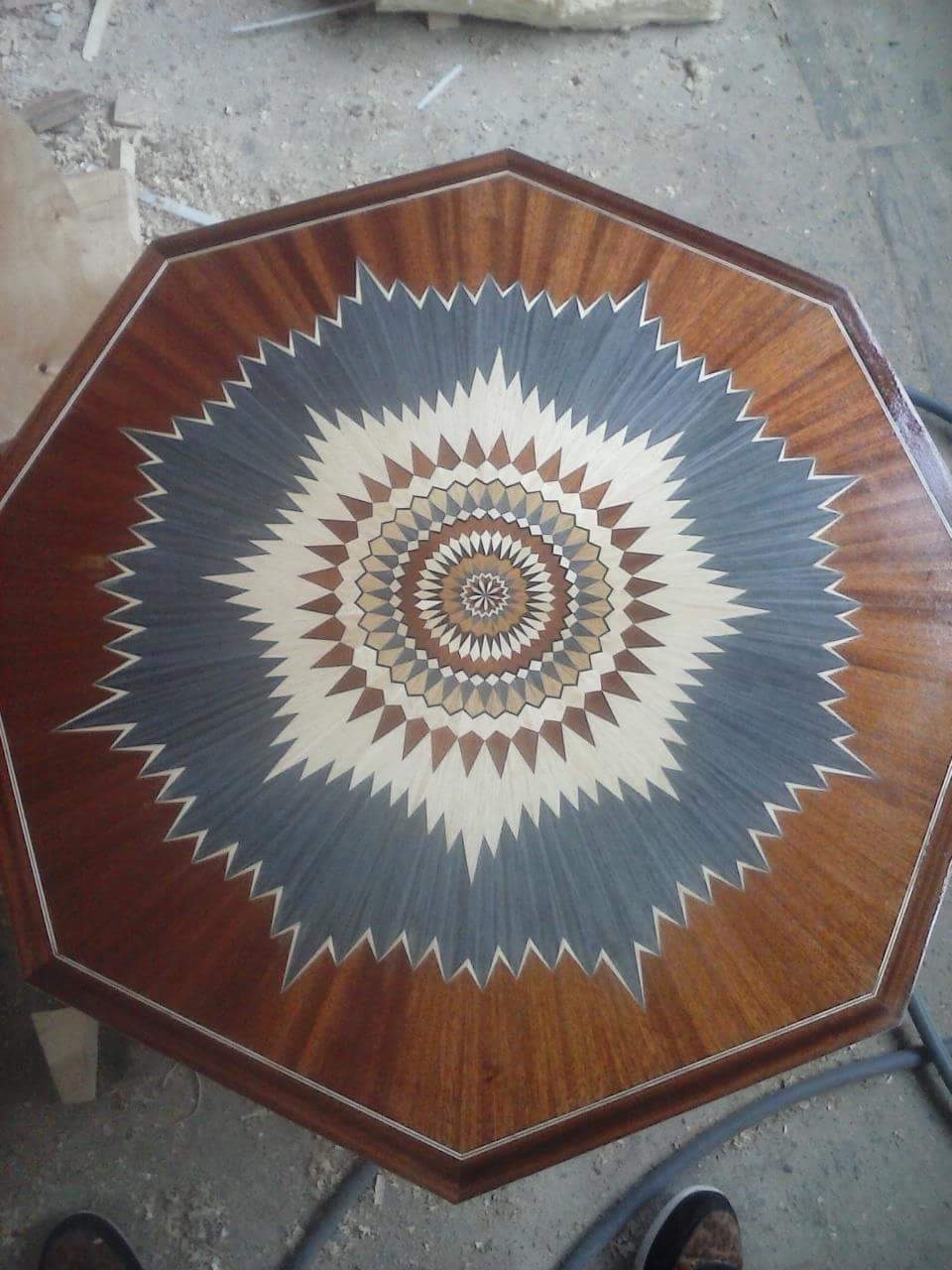 Ośmiokątny blat stołu z dekoracyjnym wzorem intarsji w odcieniach brązu, beżu, szarości i bieli, przypominającym rozetę lub kwiat.