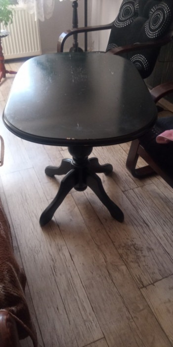 Okrągły, ciemny stolik kawowy z trzema nogami w stylu retro, ustawiony na panelach podłogowych. Widoczny fragment krzesła z ciemnym drewnem i poduszką w tle.