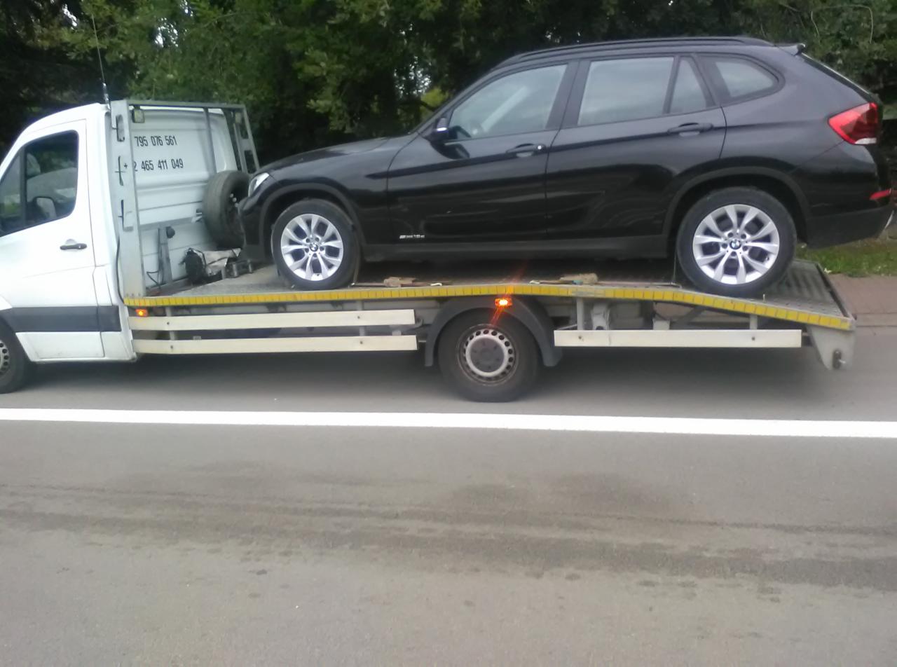 Czarny samochód BMW X1 załadowany na platformę lawety, widok z boku, biała linia na asfalcie.