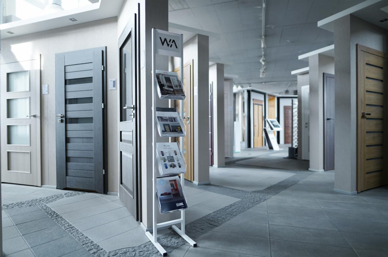 Ekspozycja salonu WAWRUK z różnymi modelami drzwi wewnętrznych i zewnętrznych, widoczne drzwi z przeszkleniami i panele drzwiowe w różnych odcieniach szarości i drewna, na pierwszym planie stojak...