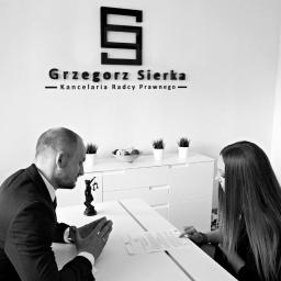 Kancelaria Radcy Prawnego Grzegorz Sierka - Spotkanie biznesowe w kancelarii, mężczyzna w garniturze i kobieta przy białym stole analizują dokumenty, w tle logo firmy i figurka Temidy.