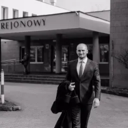 Mężczyzna w garniturze wychodzący z budynku Sądu Rejonowego, trzymający płaszcz w ręku, w czarno-białej tonacji.