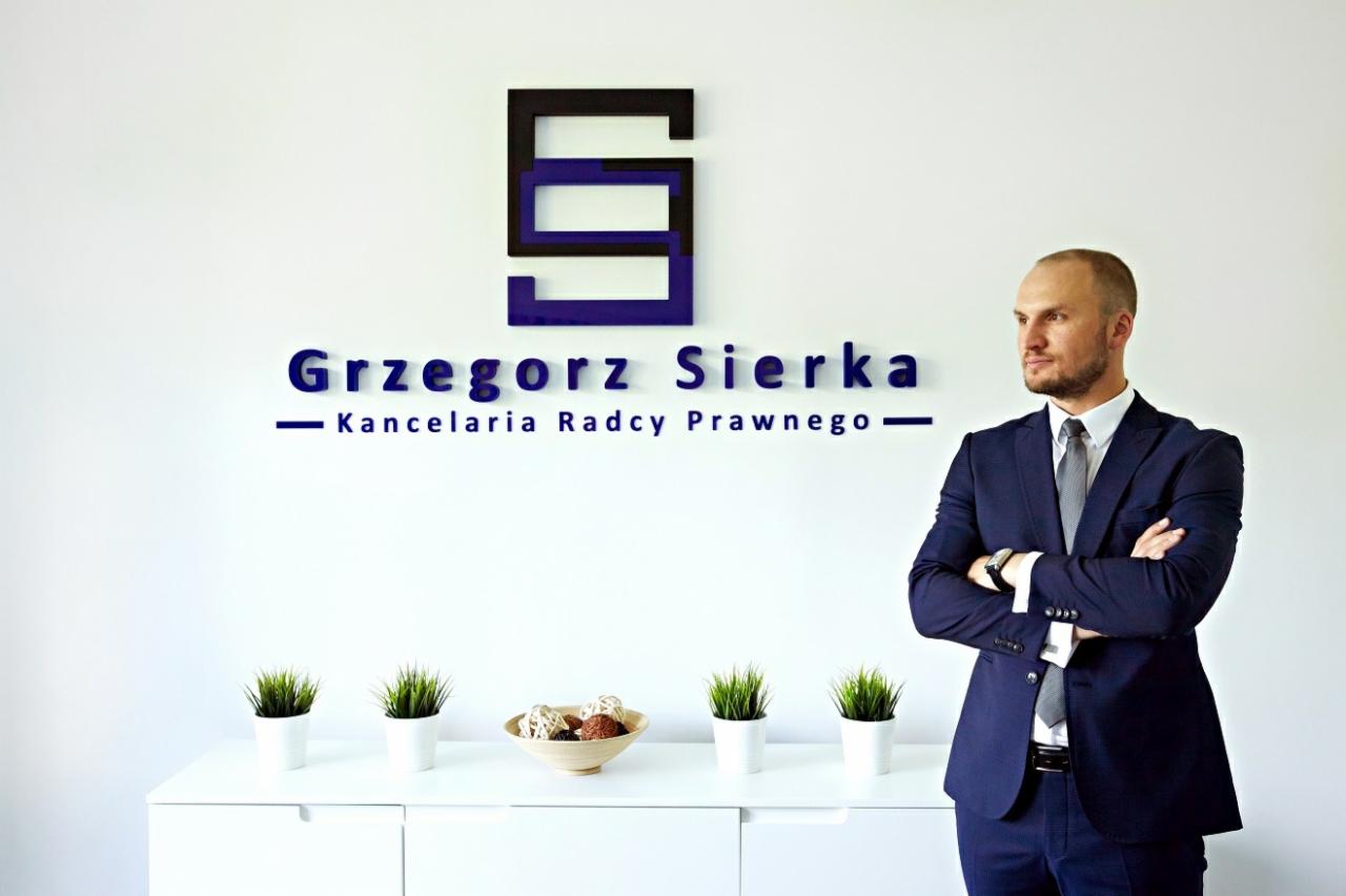 Mężczyzna w granatowym garniturze stoi na tle ściany z logo i napisem 'Grzegorz Sierka Kancelaria Radcy Prawnego', na białej komodzie doniczki z trawą i miseczka z dekoracyjnymi kulami.