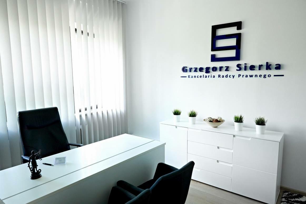 Nowoczesne biuro: białe meble, minimalistyczny wystrój, logo kancelarii 'Grzegorz Sierka' na ścianie, figura Temidy na biurku.