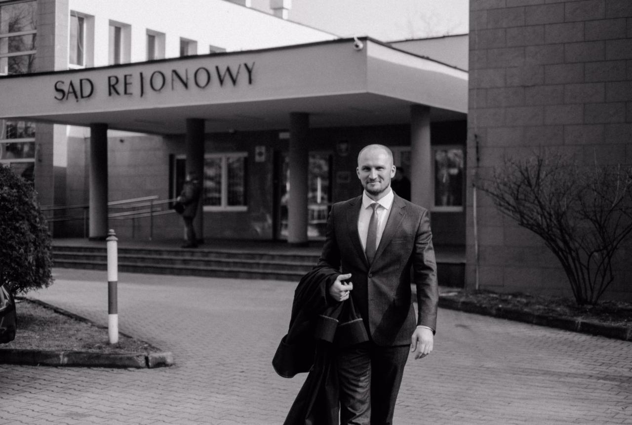 Mężczyzna w garniturze wychodzący z budynku Sądu Rejonowego, trzymający płaszcz w ręku, w czarno-białej tonacji.