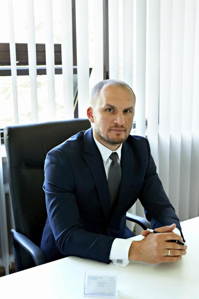 Portret mężczyzny w garniturze siedzącego za biurkiem z wizytówką w przezroczystym pudełku, żaluzje w tle.