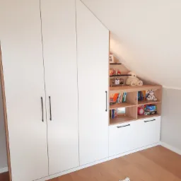 Biała szafa wnękowa z drewnianymi akcentami, dopasowana do skosu dachu, z otwartymi półkami wypełnionymi książkami i zabawkami.