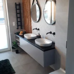 Nowoczesna łazienka z dwoma owalnymi umywalkami nablatowymi, czarną armaturą, okrągłymi lustrami w czarnych ramach, drewnianą ścianką działową i szarą szafką pod umywalkami.