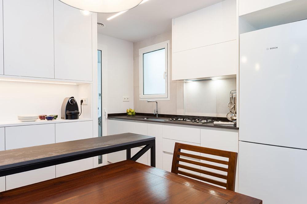 Nowoczesna kuchnia z białymi, minimalistycznymi szafkami, ciemnym blatem i drewnianym stołem z krzesłem. Widoczny ekspres do kawy i owoce w misce.