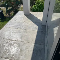 Szara, betonowa podłoga balkonu z widocznymi łączeniami płyt, dwa białe filary i duże okno balkonowe z refleksami zieleni z zewnątrz.