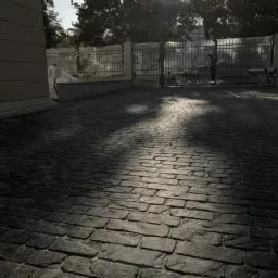 Ciemnoszara kostka brukowa ułożona na podjeździe, z widocznymi cieniami drzew i budynkiem w tle.