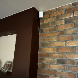Biały czujnik alarmowy zamontowany na ceglanej ścianie w rogu pokoju, widok z dołu, obok brązowa ściana z oświetleniem punktowym.