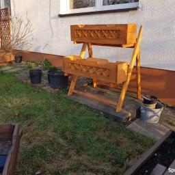 GREG-STOL Mała Stolarnia - Dwupoziomowy, drewniany kwietnik z ozdobnym ażurem w kształcie krzyżyków, ustawiony na trawniku przed białym domem z brązową podmurówką, obok doniczki i wiadro z ziemią.