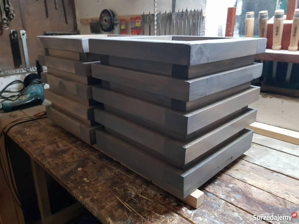 Stos szarych, drewnianych ram na roboczym stole w warsztacie, widoczne narzędzia i ślady pracy.