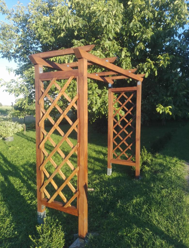 Brązowa, drewniana konstrukcja z kratownicą, prawdopodobnie wejście do ogrodu, z widocznymi metalowymi elementami łączeniowymi, na tle zielonego trawnika i drzew.