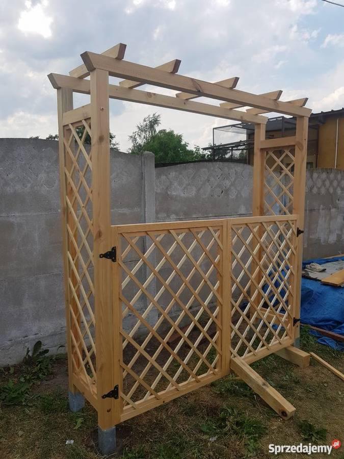 Surowa drewniana konstrukcja z kratownicą, prawdopodobnie wejście do ogrodu, jeszcze nie wykończona, z widocznymi elementami podtrzymującymi i betonowymi podstawami.