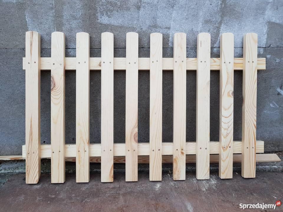 Fragment surowej, drewnianej konstrukcji ogrodzenia z jasnego drewna, z widocznymi śladami po wkrętach, ustawiony na szarej powierzchni, na tle betonowej ściany.